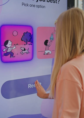 Egy szőke hajú lány keze egy interaktív kijelzőn lévő "Restart" gomb felé nyúl. A kijelzőn két grafikus opció látható: az egyiken egy fiú kutyával fut, a másikon egy fiú rakétával játszik. Interaktív kijelző, érintőképernyő, játék, választás, tanulás, dig