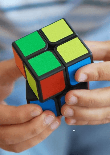 Közelkép két kézről, amelyek egy 2x2-es Rubik-kockát tartanak és forgatnak. A kocka színes oldalai (zöld, sárga, kék, piros, narancs) jól láthatók. A háttérben egy kék-fehér csíkos ing látható, elmosódva. Logikai játék, Rubik-kocka, fejtörő, intelligencia